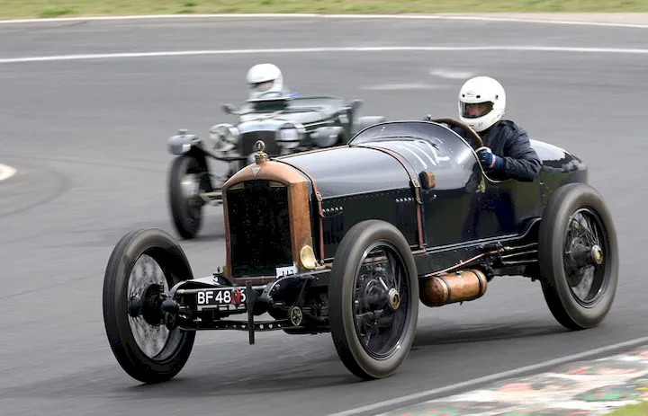 An example of an open-top race car from the early 20th century. The drivers' helmets are modern.